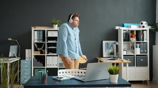 Slow motion of funny employee attractive guy dancing in office room wearing headphones having fun alone. Modern gadgets, joy and entertainment concept.