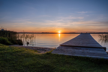 Colorful orange sunset on a quiet lake. Perspective view of a wooden pier on the pond at sunset.