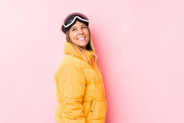 Young caucasian woman wearing a ski clothes in a pink background looks aside smiling, cheerful and pleasant.