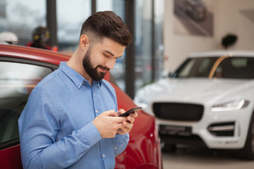 Attractive bearded man smiling whle using his smart phone at car dealership, copy space. Texting,...