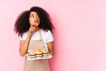 Young afro pastry maker woman holding a cupcakes isolatedYoung afro baker woman looking sideways with doubtful and skeptical expression.