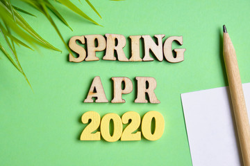 The concept of spring - April in the new year. Wooden numbers 2020 with letters and sheet of paper with a pencil, leaves on a green background. Top view.