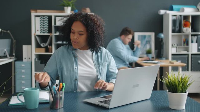 African American young woman office worker is busy using modern laptop taking notes at work at desk while male coworker is working in background.