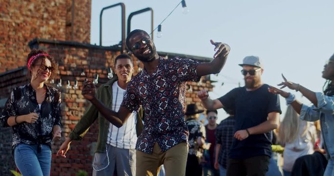 Cheerful young and stylish African American guy dancing cool in the middle of his multiethnic friends on the rooftop at party. Outdoors.
