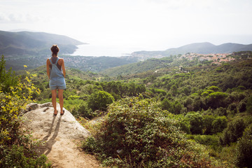 Young woman admiring panorama from hill overseeing bay in Tuscany wearing denim dungaree