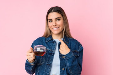 Young caucasian woman holding a tea cup pointing with finger at you as if inviting come closer.