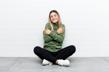 Young caucasian woman sitting on the floor hugs, smiling carefree and happy.