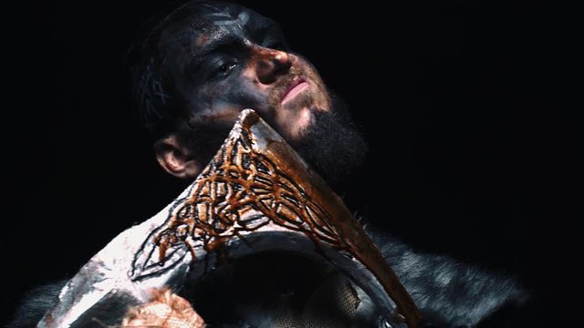 Viking holds an ax blade along the bristles. Warrior in a combat color with an ax near his cheek. Fiery red background