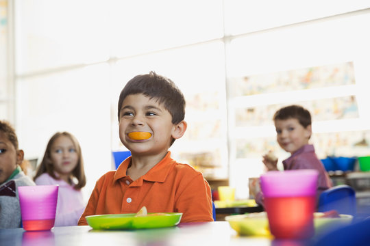 Playful Boy Holding Orange Slice In Mouth At School At Snack Time