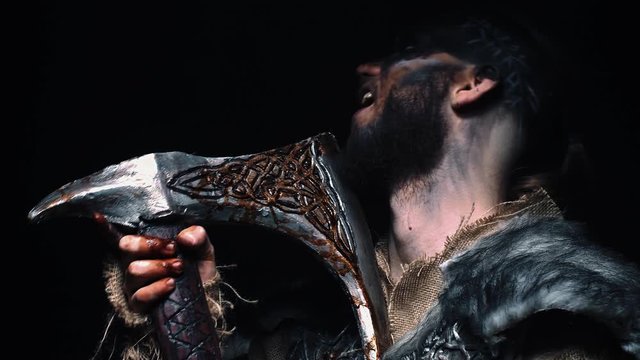 The Viking man looks into the camera and slowly lowers his head, the warrior holding the axe next to his face. Viking in war paint