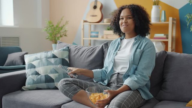 Slow motion of pretty Afro-American lady laughing watching TV eating snacks enjoying funny show at home. Entertainment, people and house concept.
