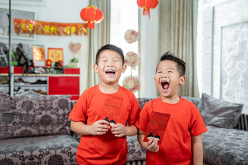 two kids smile in red shirts with angpau standing at front a couch - Powered by Adobe