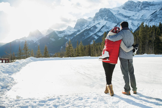 Couple with ice skates hugging below snowy mountain