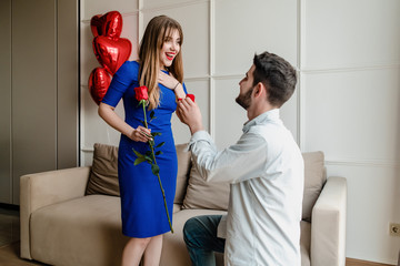 man proposes to woman with ring and red rose at home with heart shaped balloons