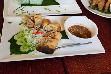 Fried spring rolls with with sliced cucumber and sweet chili sauce in a white plate on red table, Vietnamese food in Cambodia