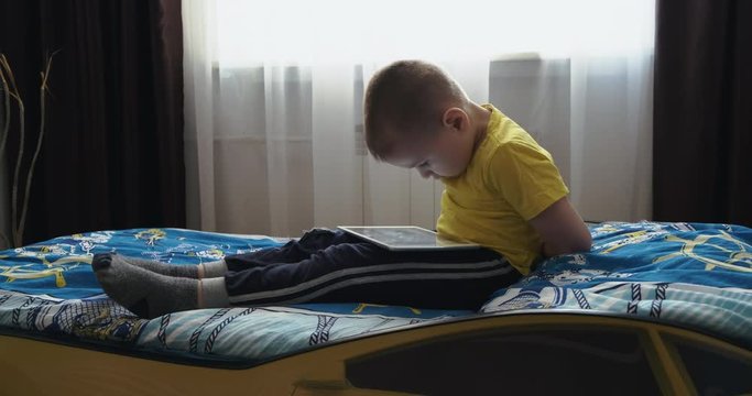 Five-year-old boy sits on bed in his room, in home clothes, yellow T-shirt, tablet lies on his lap, child is hunched over looking at screen uninterrupted, cartoons, games, new technologies in training