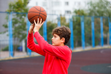 Cute boy in red t shirt plays basketball on city playground. Active teen enjoying outdoor game with orange ball. Hobby, active lifestyle, sport for kids.