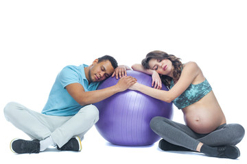 Fototapeta na wymiar A young couple and their wife in the last months of pregnancy are sitting on a sports mat near the ball. Black man and white woman. The concept of a happy couple of different races preparing together