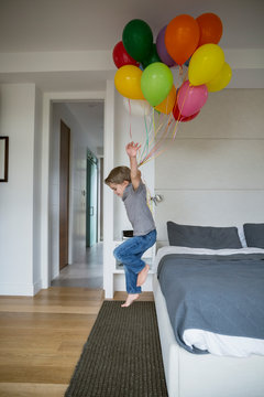 Boy with bunch of multicolor balloons jumping off bed