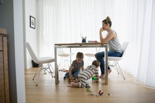 Brothers playing with toys under mother working at laptop