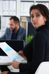 young beautiful woman sit on chair at table in office in cabinet of her boss hold binder in arms work with documents