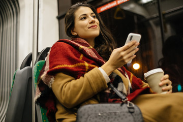 Portrait of a young woman at the public transport, sitting with coffee and using her cell phone  in...