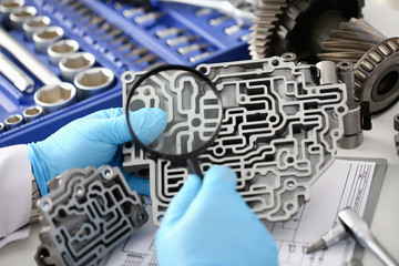 Auto service repairman for automatic gearboxes keeps in hand in blue protective gloves examines through the lens the detail of the hydraulic unit performs diagnostics and estimates the detail closeup.