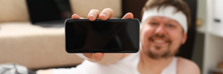 A young cute fat man with overweight does the first fitness workout at home demonstrates a smartphone in the camera with a training program holds in his hand.