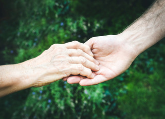 Fototapeta premium Senior and young hands outside closeup. Elderly woman and young man holding hands together, green blurred background, sunlight. Love, warmth, take care concept, Valentines, mothers day, donate, help.