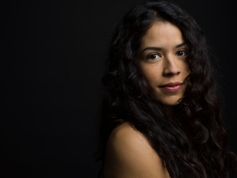 Portrait confident Latina woman with bare shoulders and long curly black hair against black background