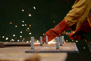 Defocused of arc welder hand wearing red safety welding glove performing welding steel bolts into the metal plate at the construction fabrication site  workshop 