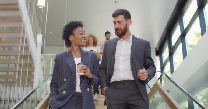 Multiethnic colleguaes discussing business and communicating to each other while descending modern office stairs. Man and woman walking downstairs and talking with coworkers at back.