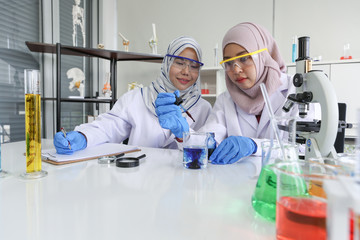 Young Asian muslim female scientist wearing safety glasses holding blue reagent dropper with beaker in laboratory room