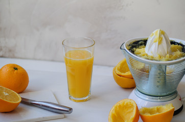 On a wooden table, one whole orange, one cut in half, peel of an orange. On the table lies a stone board and a knife. In the background is a glass cup with freshly squeezed orange juice. On the right