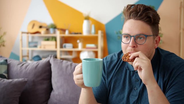 Portrait Of Cheerful Young Man Drinking Coffee And Eating Biscuit Sitting On Couch Alone In Apartment Enjoying Snack. Food, Drinks And Leisure Time Concept.