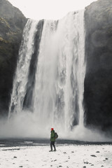 young male traveler in green clothes in a red hat and a green backpack stands near a waterfall in Iceland