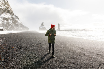 young male traveler in green clothes in a red hat and a green backpack goes black beach in Iceland