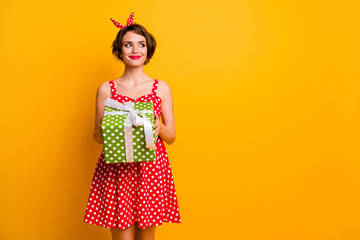 Photo of pretty lady hold large green giftbox look side empty space tricky excited birthday girl wear retro style summer dotted red white dress headband isolated yellow color background