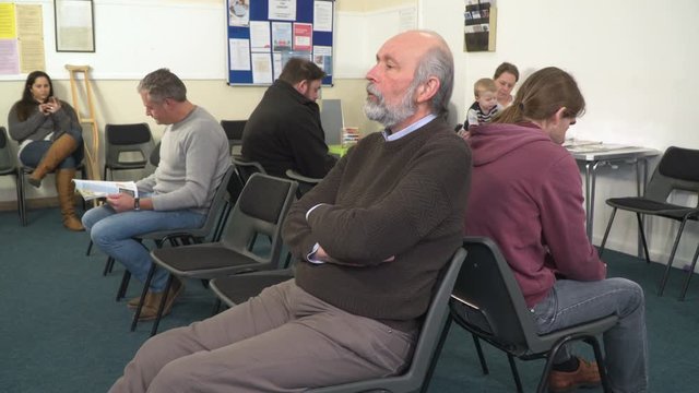 4K: People sat in Waiting room awaiting Healthcare appointments. Could be a Doctor or dentist surgery clinic. Man sighing