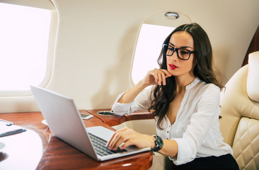 A nice businesswoman in formal attire and glasses, who is typing something on her laptop with thoughtful face expression, touching her chin while flying on a private jet.