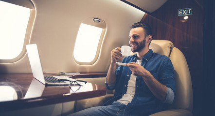 Luxury break. Young cheerful man in denim outfit is smiling while drinking coffee, sitting in a window seat near the desk with a laptop on it while flying on a private jet.