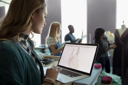 Female fashion design student using laptop, viewing dress designs in studio