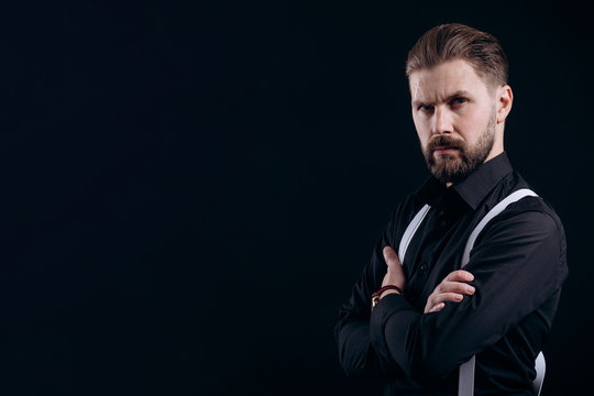 Half-turned portrait of masculine bearded man in black shirt and white suspenders isolated black background copyspace