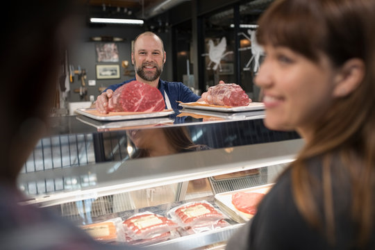 Butcher Showing Raw Meat Cuts To Customers At Display Case In Butcher