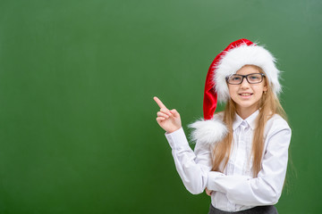 Smiling girl wearing a red santa hat and eyeglasses points with finger away on empty chalkboard. Space for text