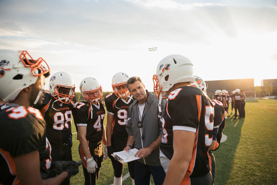 Coach with clipboard talking to teenage boy high school football team in huddle on football field