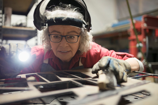 Senior female machinist with flashlight using laser cutter, examining laser cut wood in workshop