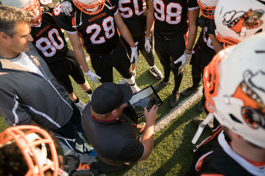 Coach with digital tablet showing video to teenage boy high school football team in huddle on football field
