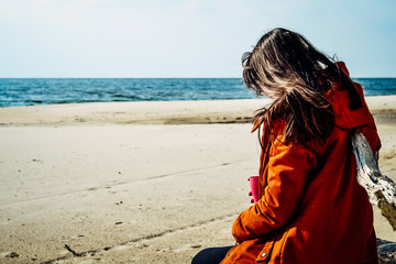 young beautiful caucasian girl in a red warm jacket with a hood, scarf, leather tight pants sitting on a rocky beach resting after a walk by the sea