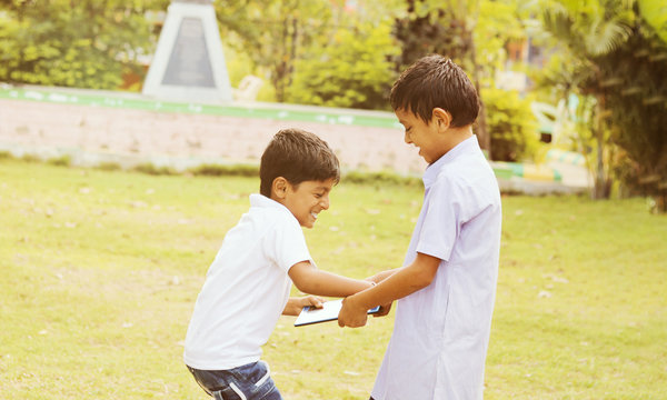 Two kids fighting for tablet at park outdoor - Liite boy trying to grab a tablet from her friend or brother - Concept of children's addiction, aggression on electronic devices or gadgets. - Powered by Adobe